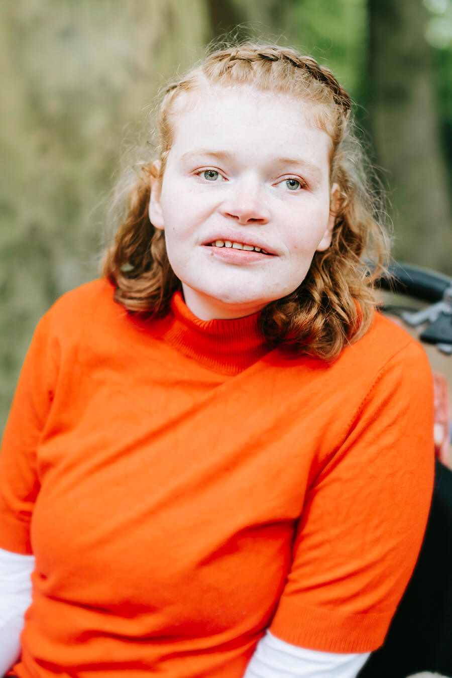 Elfi kijkt recht in de camera. Ze draagt een feloranje trui en zit in haar rolstoel in het bos.