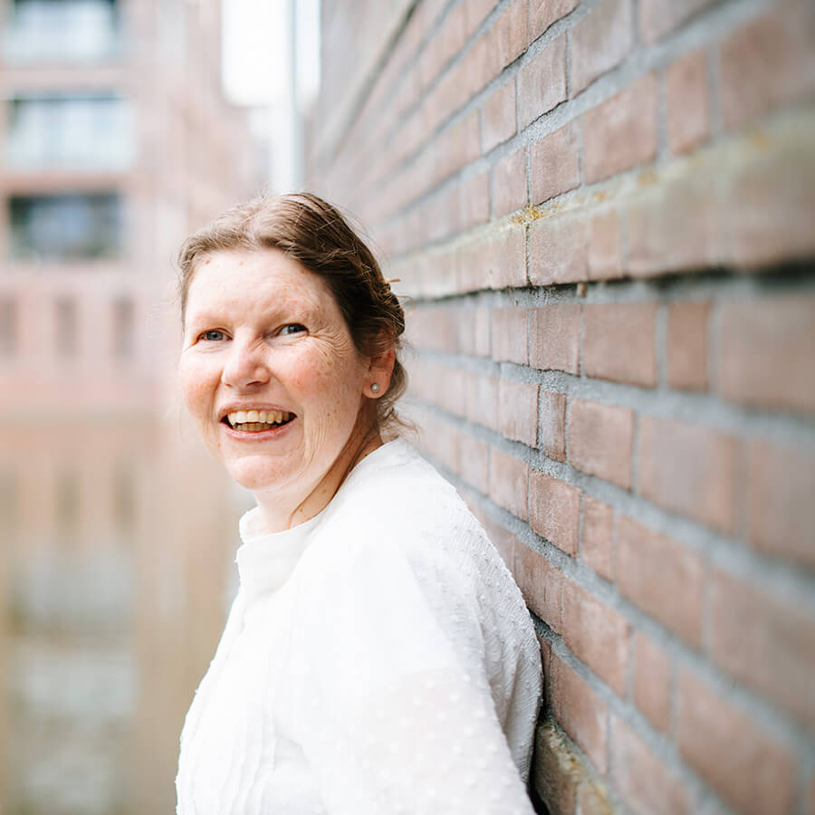 Juline staat zijwaarts tegen een muur en kijkt lachend over haar schouder naar de camera.